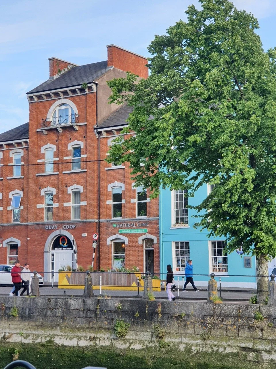 The Natural Clinic Street view with red brick and light blue buildings, a large green tree, pedestrians walking, and a clear sky.