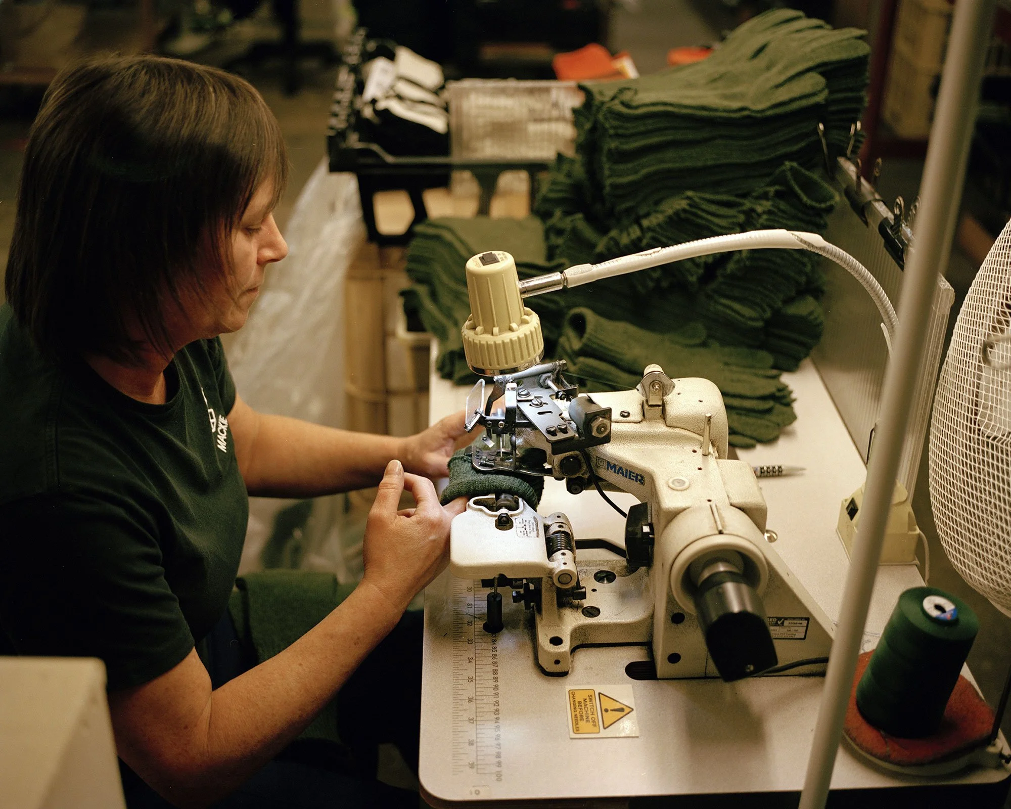 Factory worker working with a sewing machine in a knitwear mill with neatly stacked green knitted pieces on a table.