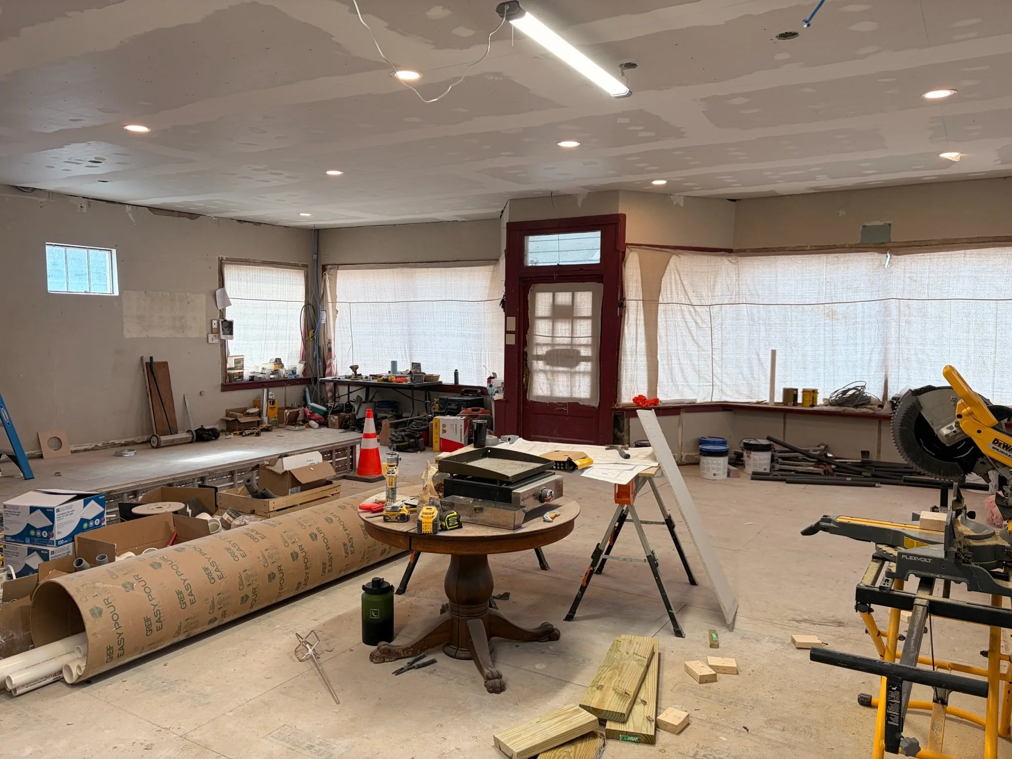A room under renovation with tools, construction materials, and equipment scattered around. The ceiling is unfinished with exposed drywall and installed recessed lighting. Large windows covered with protective sheets are visible, along with various construction supplies and a circular saw on the right side.