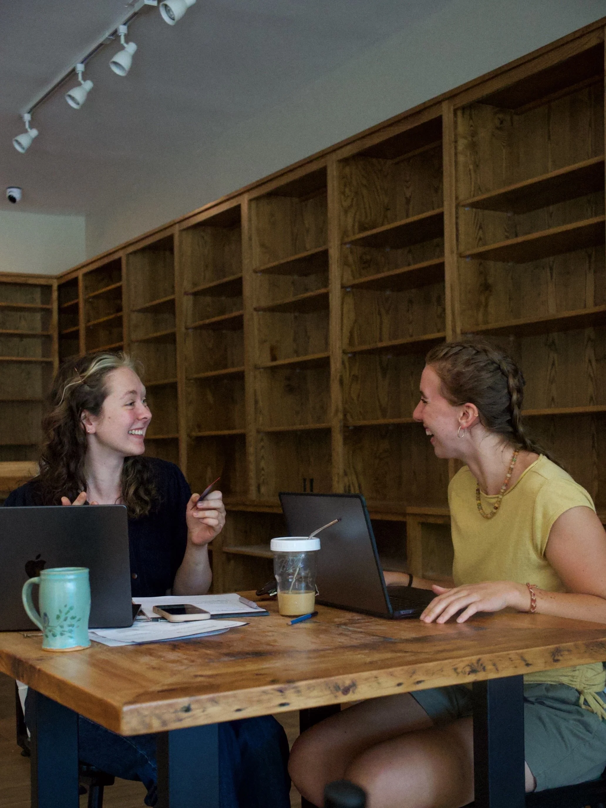 Two women sitting at a wooden table in a room with wooden shelves and ceiling track lights, engaged in conversation and smiling.