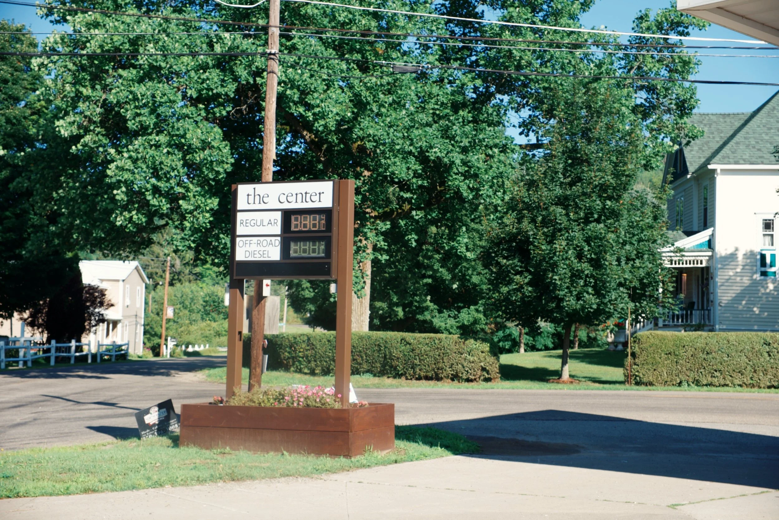 A gas station sign displaying fuel prices, located in a suburban neighborhood with trees, houses, and a clear blue sky.