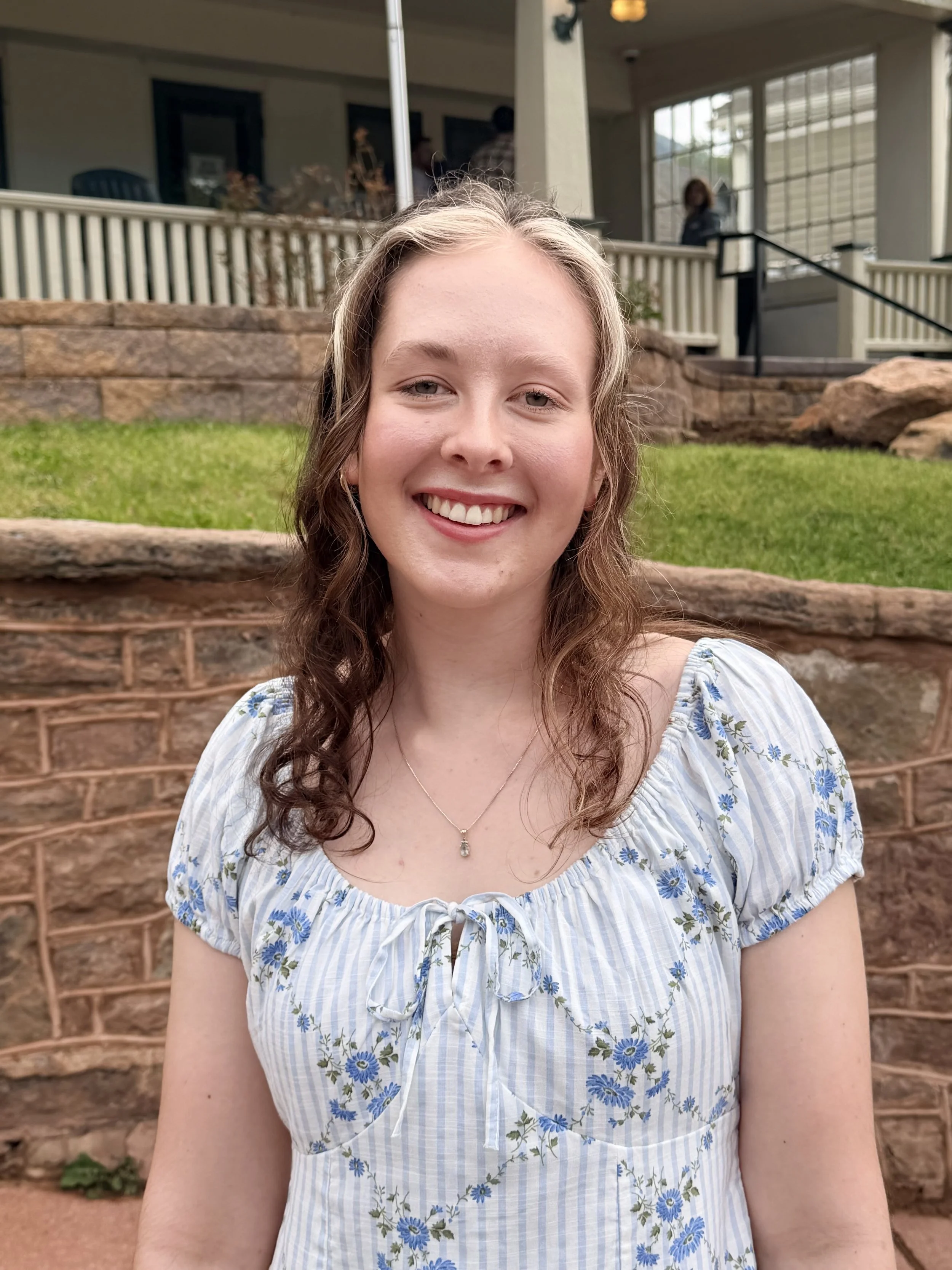 A young woman with light skin and curly, shoulder-length hair is smiling outdoors in front of a brick wall, a grassy area, and a porch with a railing. She is wearing a blue and white striped dress with floral patterns and a necklace with a pendant.