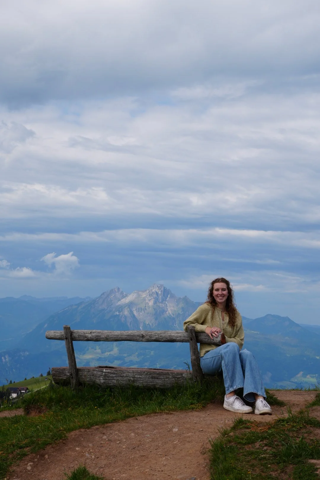 A woman sitting on a wooden bench on a mountain trail, with a mountain range and cloudy sky in the background, smiling and wearing casual clothing.