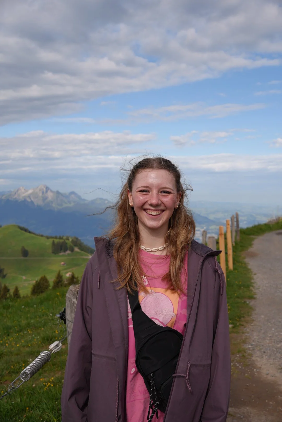 A smiling young woman with long, wavy, light brown hair standing outdoors on a mountain trail. She is wearing a purple jacket, a pink t-shirt, and a necklace made of shells. Behind her is a scenic landscape of green hills, distant mountains, and a partly cloudy sky.