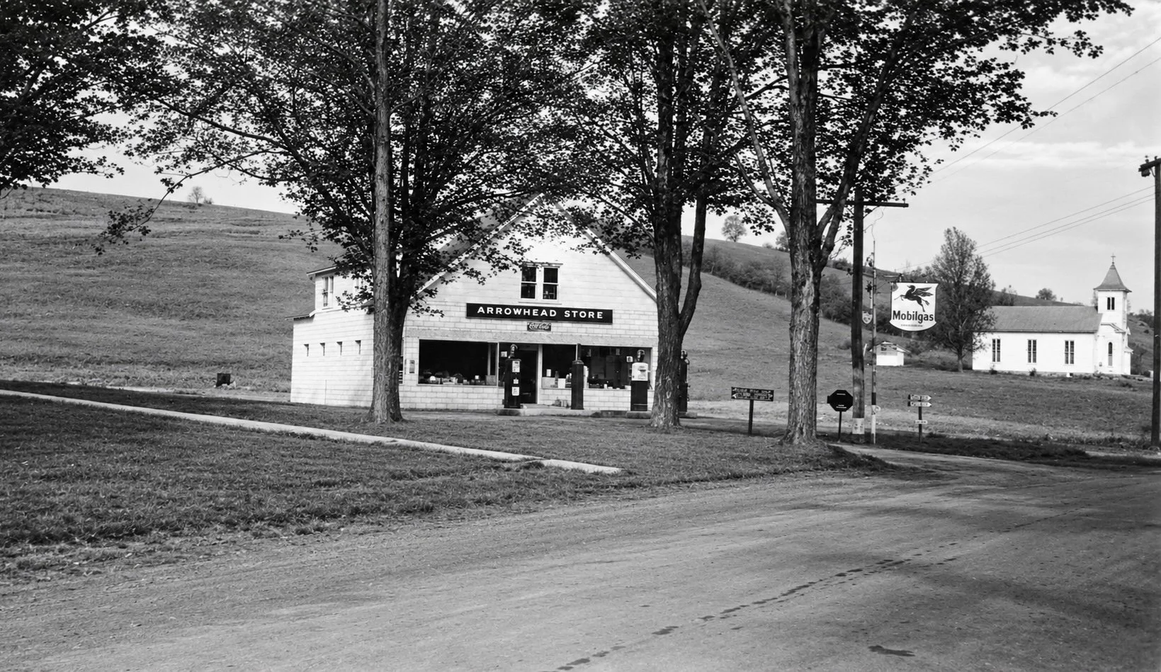 Black and white photo of a small country store called Arrowhead Store, with a sign for Coca-Cola. There are three trees in front, and a dirt road runs in front of it. In the background, there is a church with a steeple and a countryside landscape.