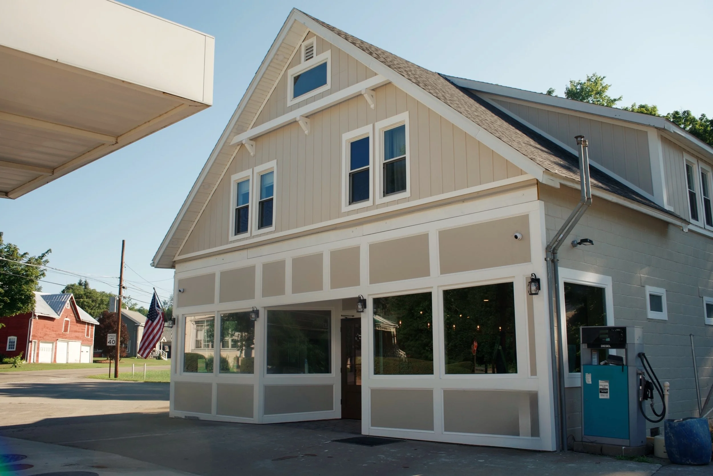 A two-story house with beige siding and white trim, featuring several windows, a steep gabled roof, and an enclosed porch area at the front. There is an American flag on a pole and a speed limit sign visible in the background. The house is situated in a neighborhood with other houses and trees, and the sky is clear and blue.