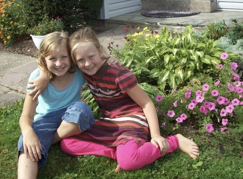 Two young girls sitting on the grass near a garden with pink flowers, smiling and hugging each other.