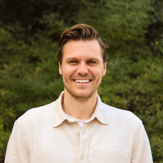 Portrait of a man with brown hair, wearing a cream shirt, standing in front of greenery smiling