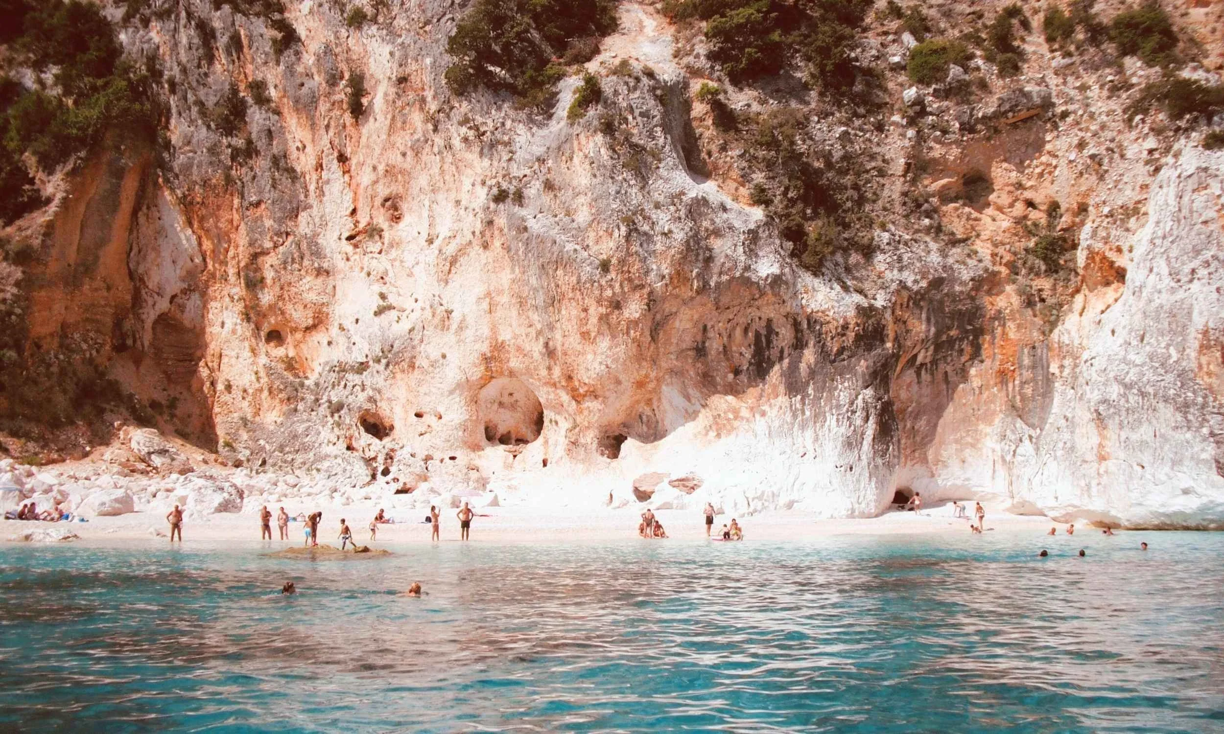 Beach scene with turquoise water and large white rocks along the shoreline, backed by towering rugged cliffs with sparse vegetation, and people swimming, sunbathing, and relaxing on the beach.