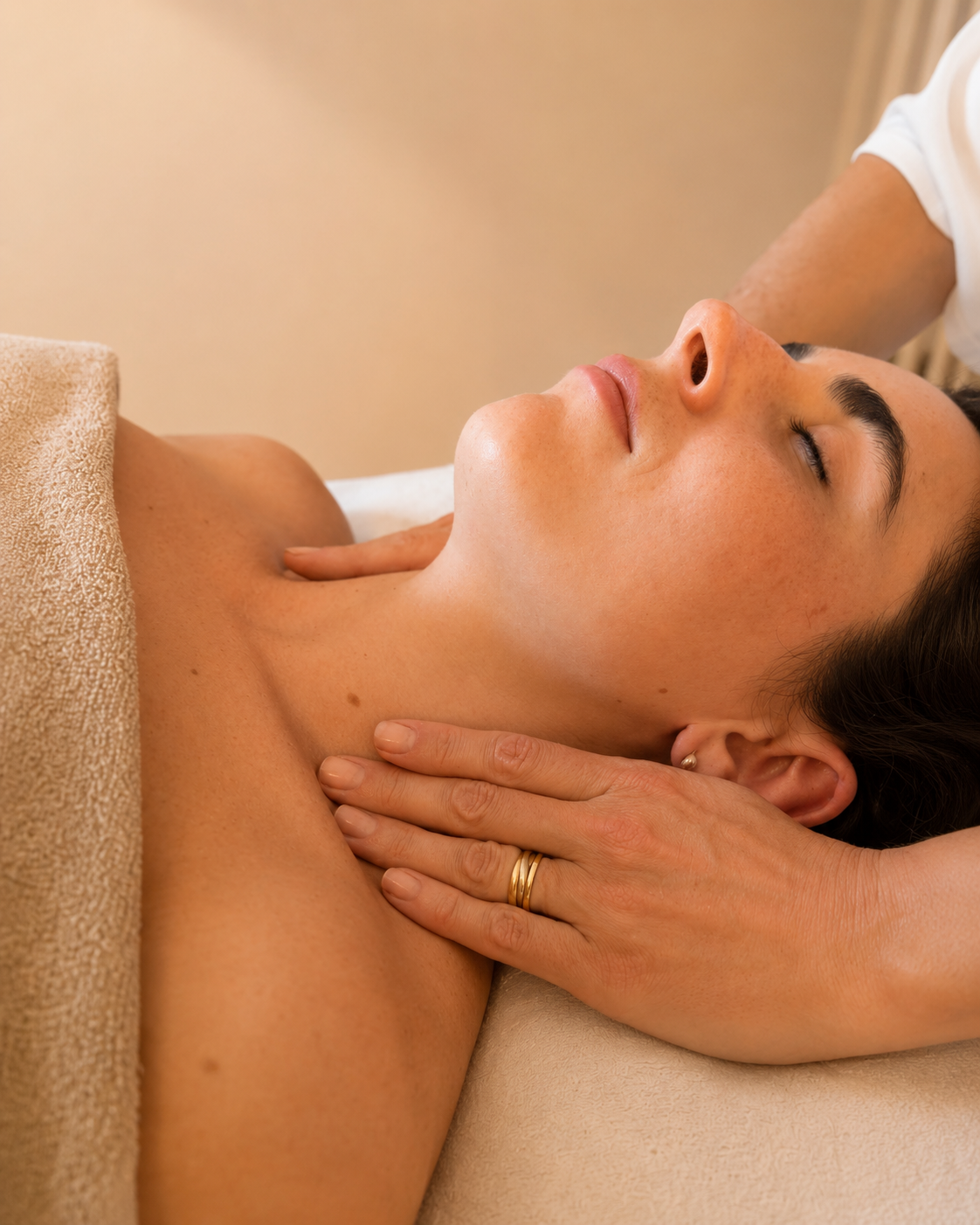 A woman receiving a massage on her shoulder and neck area, lying down with her eyes closed, covered partially with a towel, and a massage therapist's hands applying pressure.