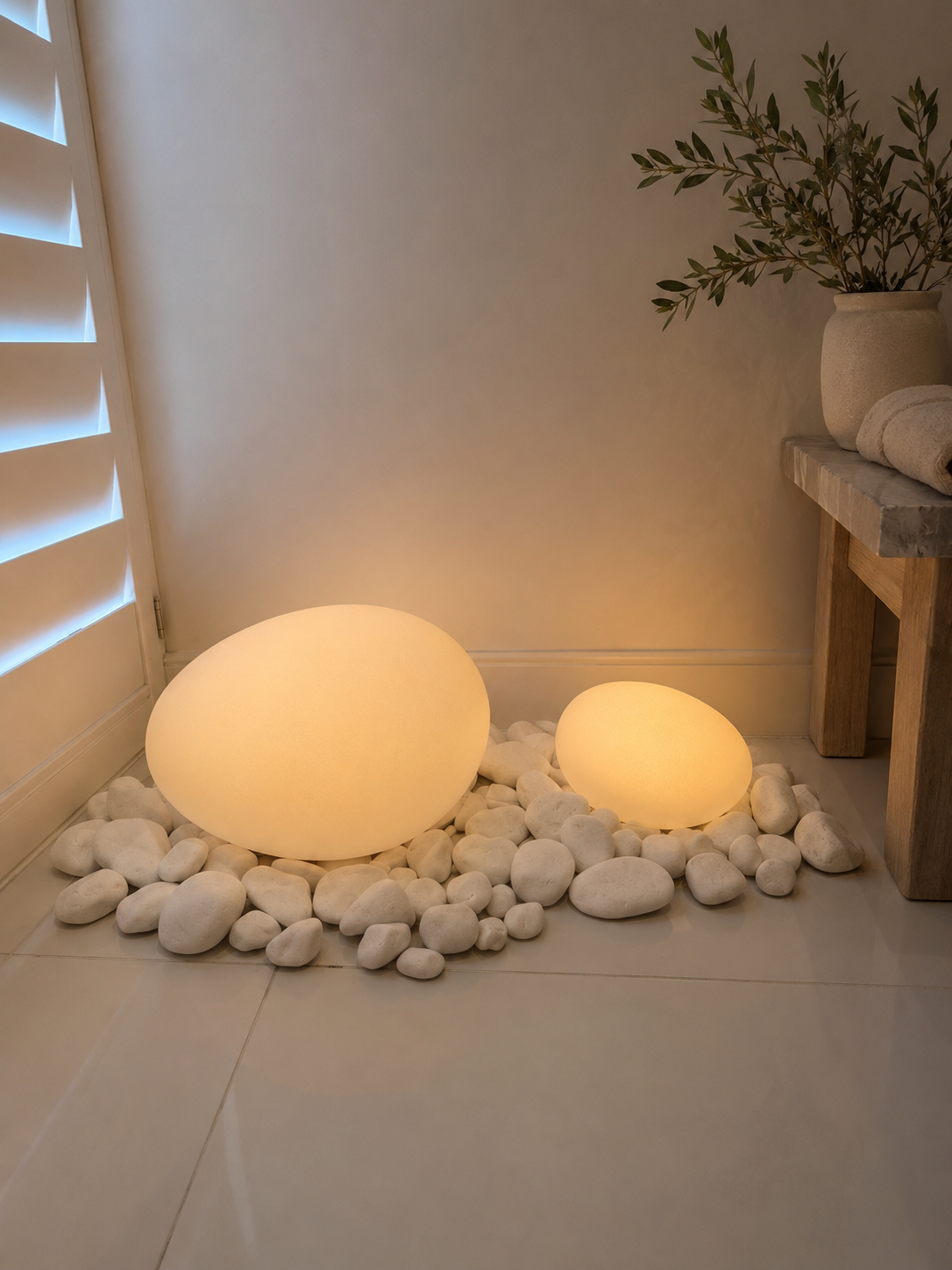 Decorative interior scene with two glowing stone-shaped lamps on white pebbles, next to a wooden table with a potted plant and towels.