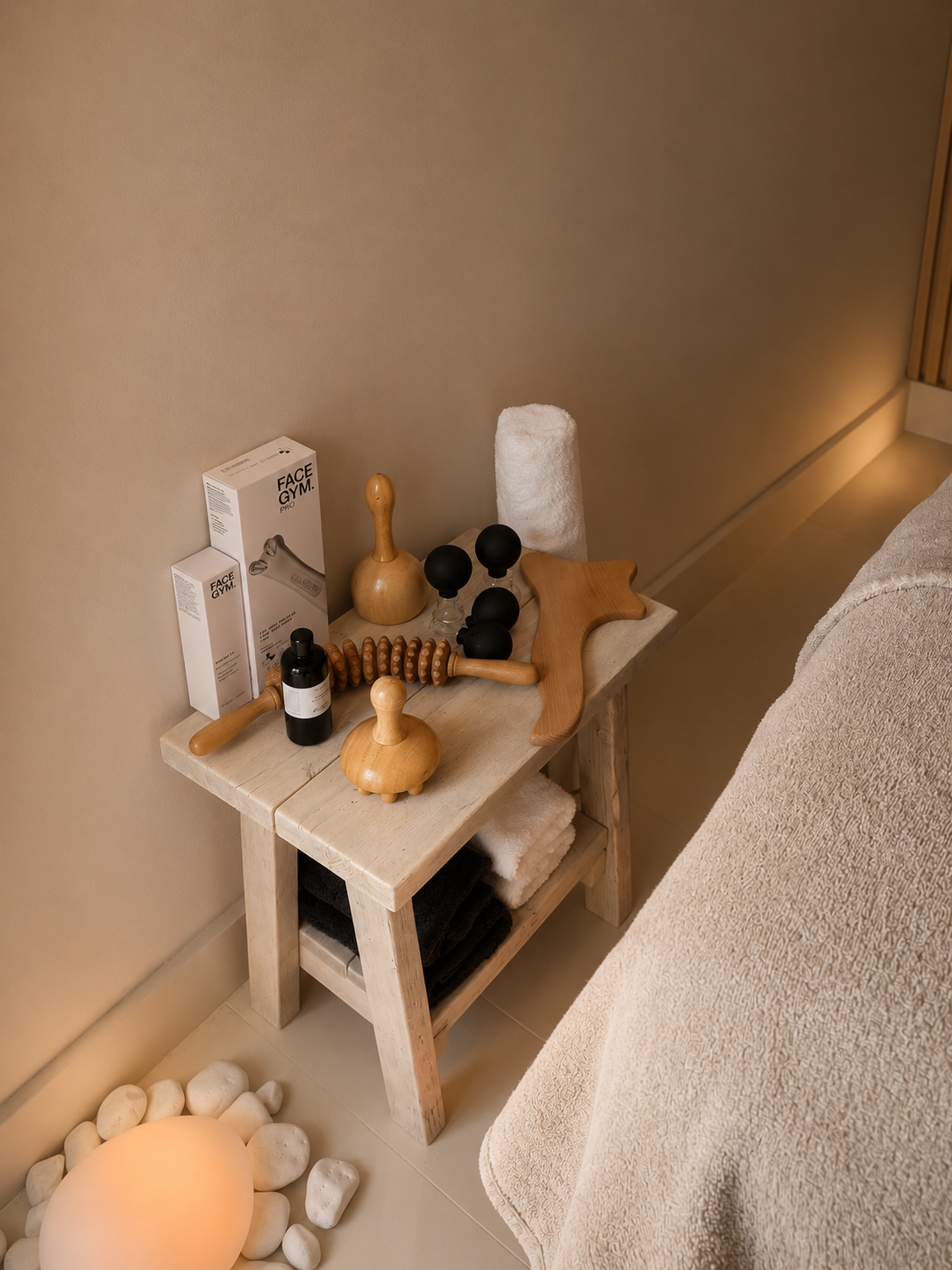 A small wooden table with massage and facial tools, including a foam roller, massage rollers, and black massage balls, along with face gym devices, placed next to a bed with beige bedding. Rocks and a spherical lamp are on the floor nearby.