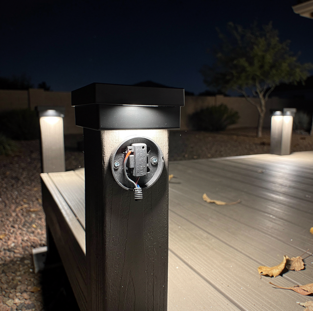 Electrical outlet mounted on black post at night with lit candles on deck near landscaping.