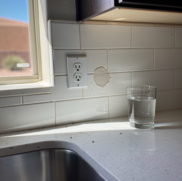 Kitchen counter with sink, white tile backsplash, electrical outlet, and glass of water near a window.