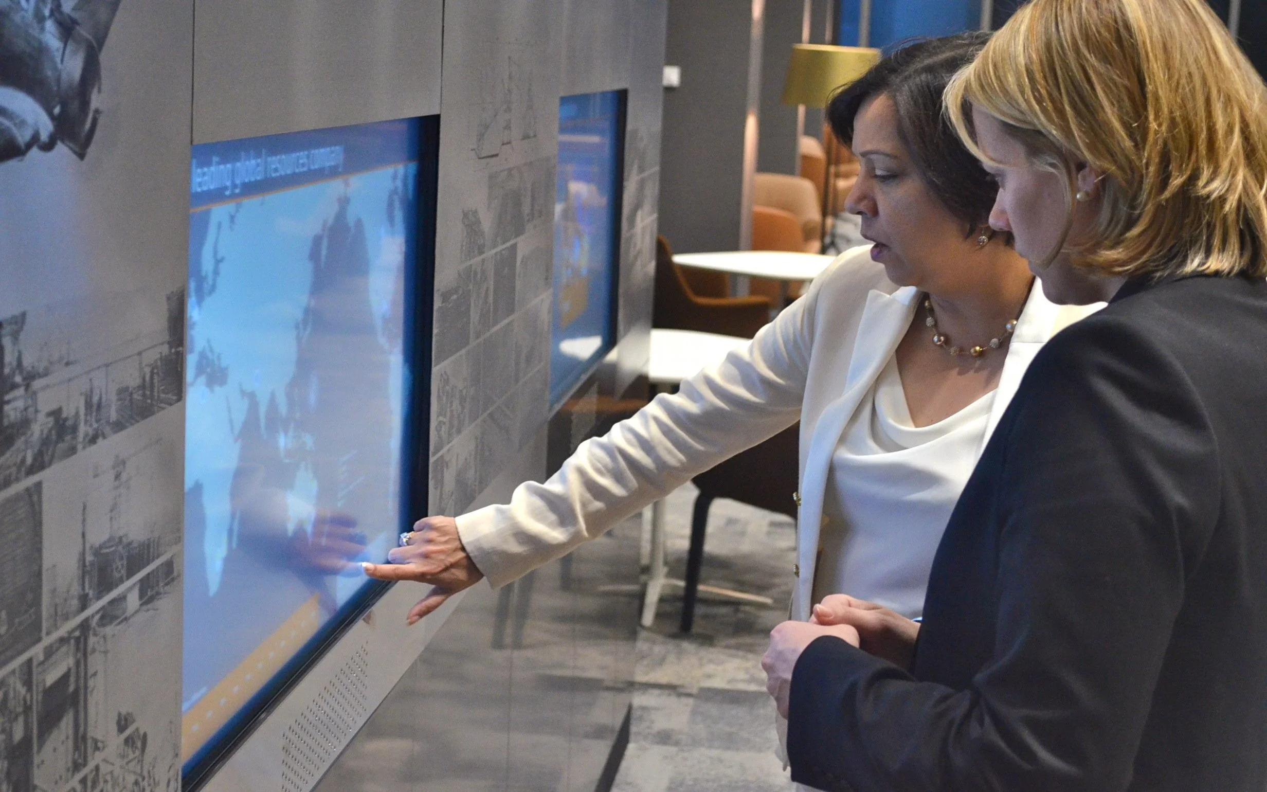 Two women in the BHP headquarters are looking at the permanent interactive display screen on a wall, with one pointing at it.