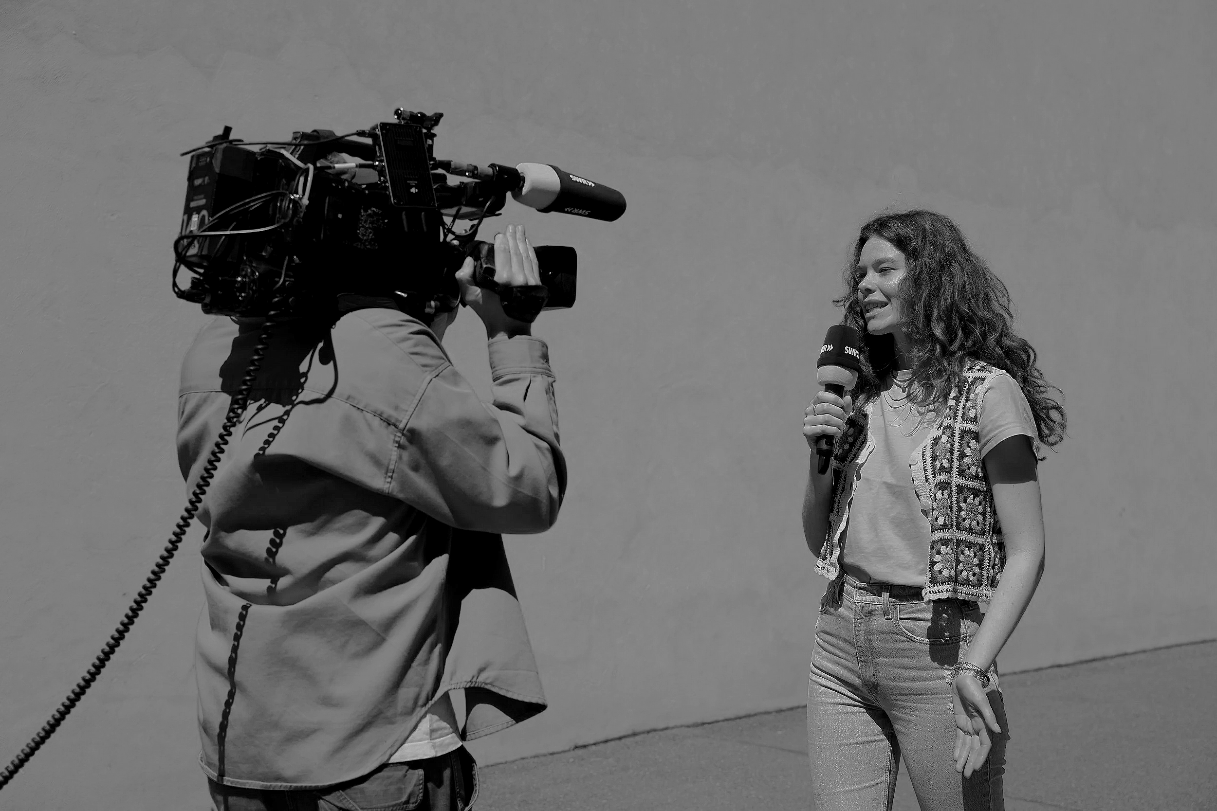 Eine Frau wird in einem Interview mit einem Mikrofon festgehalten, während ein Kameramann sie filmt, im Freien bei sonnigem Himmel.