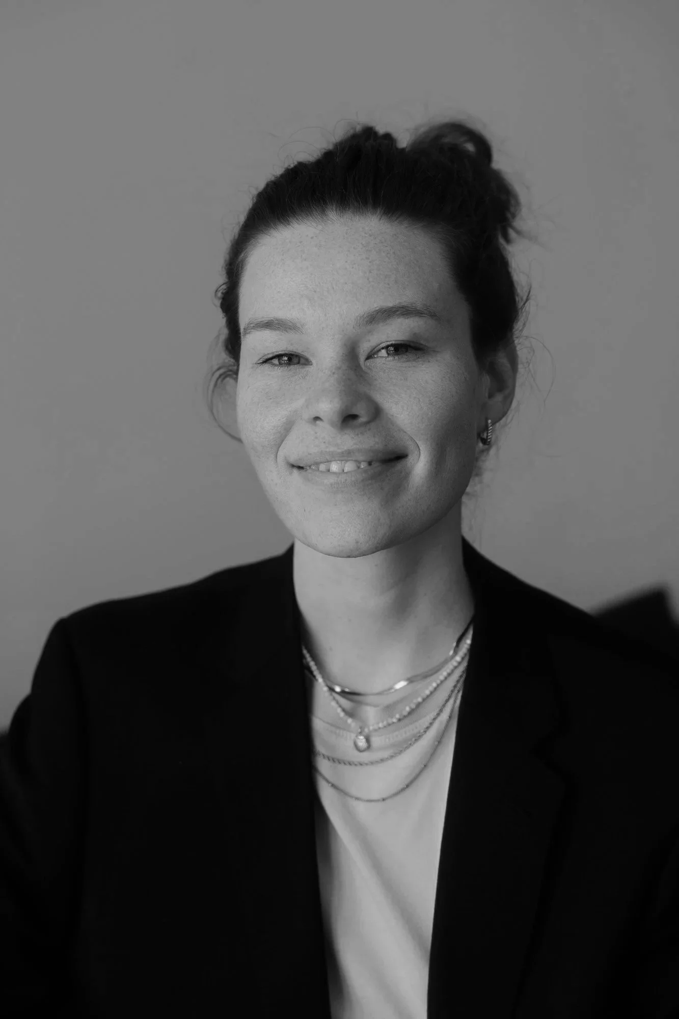Schwarz-Weiß-Fotografie eines lächelnden Frauengesichts mit geflochtenem Haar, getragenem schwarzen Blazer, mehreren Halsketten, Earcuffs und einem weißen T-Shirt vor einem grauen Hintergrund.