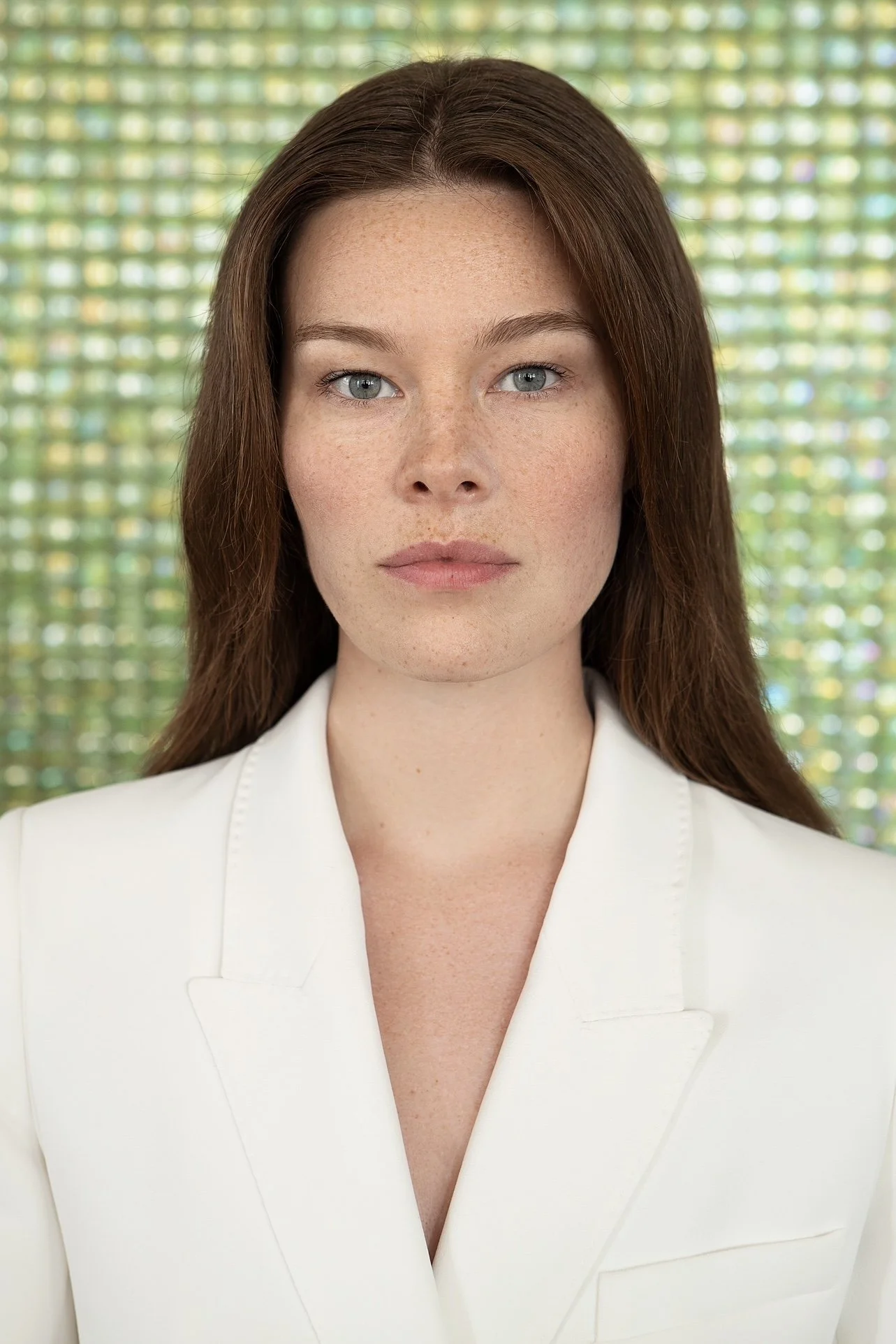 Nahaufnahme einer jungen Frau mit rotem Haar, blauen Augen und Sommersprossen, trägt weiße Kleidung, im Hintergrund eine bunte, leuchtende Wand.