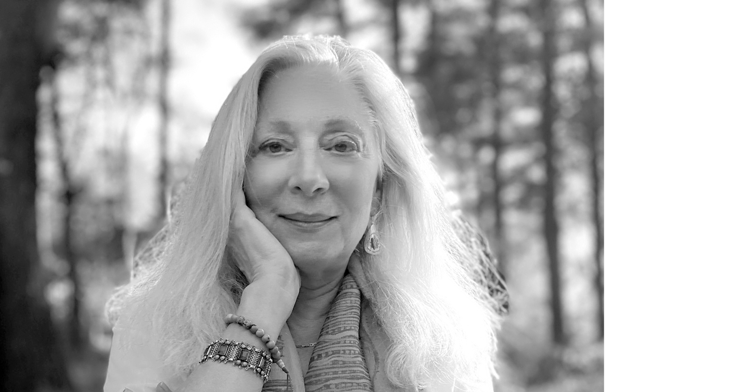A woman with long, light-colored hair smiling softly, resting her head on her hand in a forest setting.