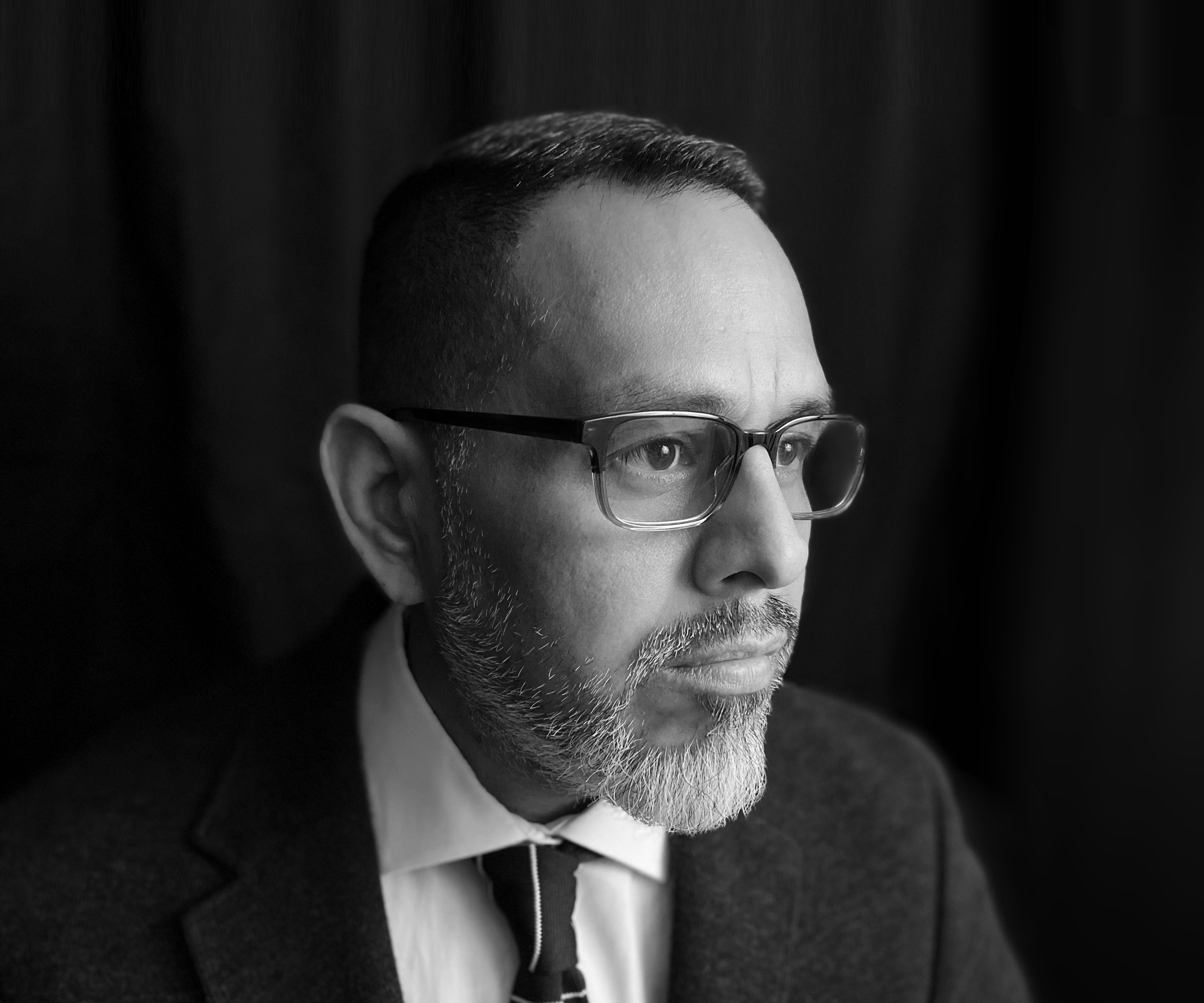 Close-up black and white portrait of a man with glasses, short dark hair, and a beard, looking thoughtfully to the right.