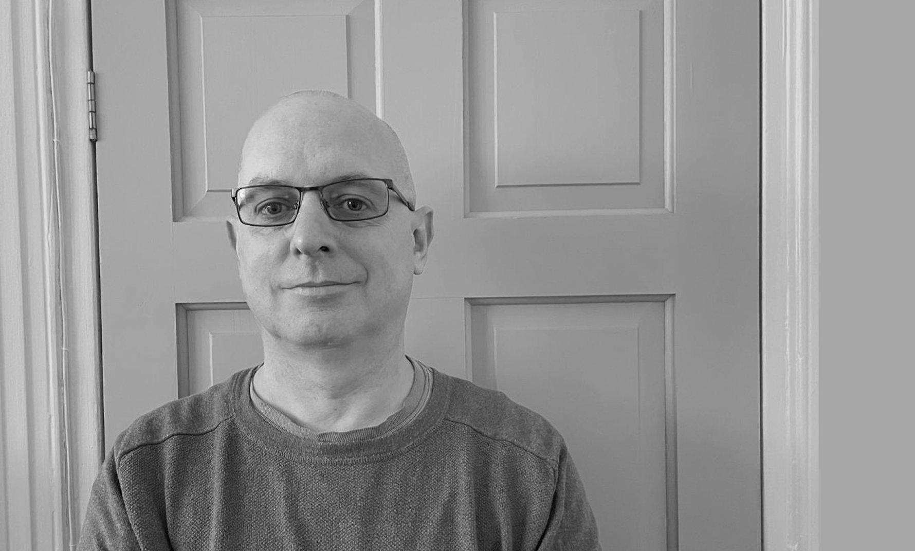 A man with glasses and a shaved head standing in front of a paneled door in a grayscale photo.