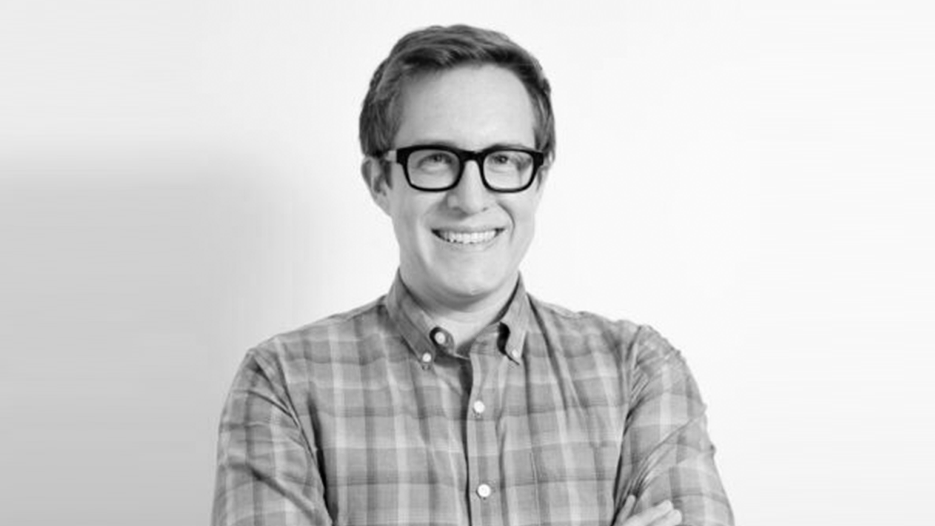 Black and white portrait of a smiling man with glasses, wearing a plaid shirt, standing against a plain background.