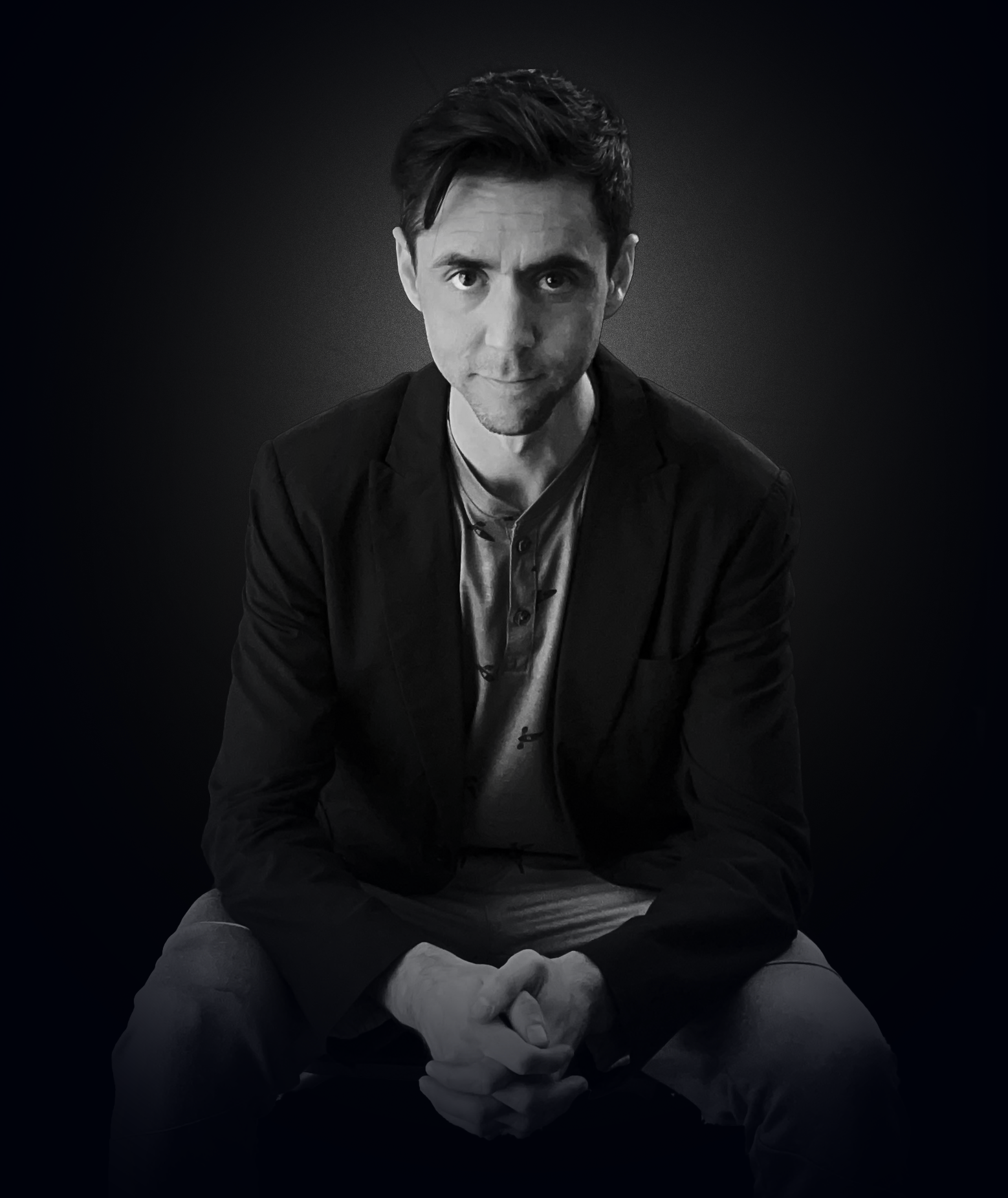 Black and white portrait of a man sitting with crossed legs, wearing a blazer over a shirt, against a dark background.