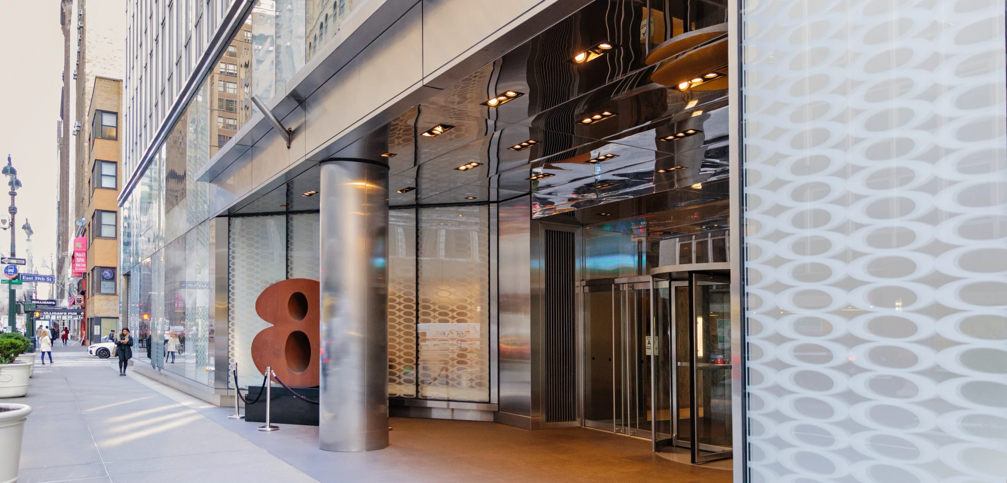 Modern building entrance with glass and metal facade, city street scene with pedestrians and street signs, large decorative orange sculpture, and a streetlamp.