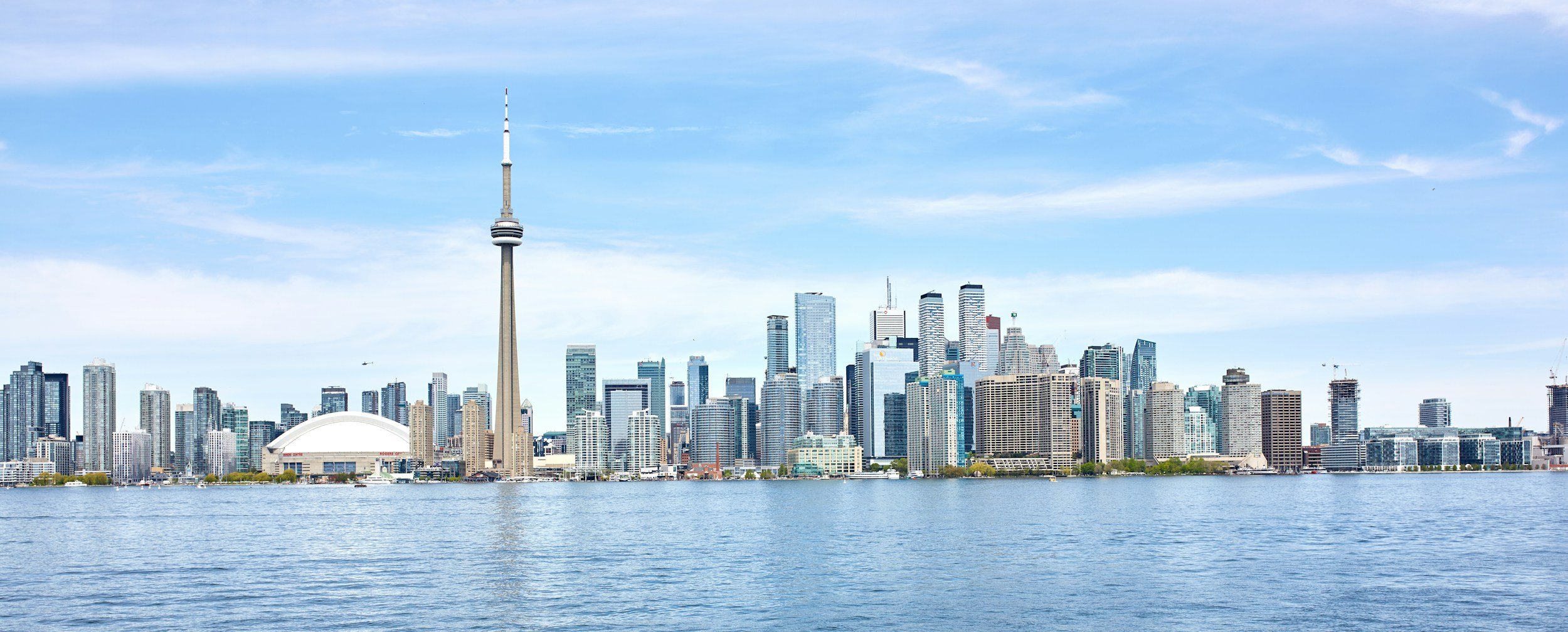 A city skyline featuring tall skyscrapers and a prominent TV tower, viewed across a body of water under a partly cloudy sky.