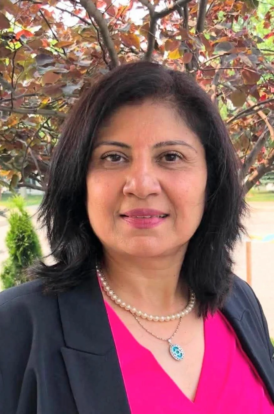 A woman with black hair wearing a pink top, black jacket, pearl necklace, and matching pendant necklace standing outdoors in front of a tree with reddish-brown leaves.