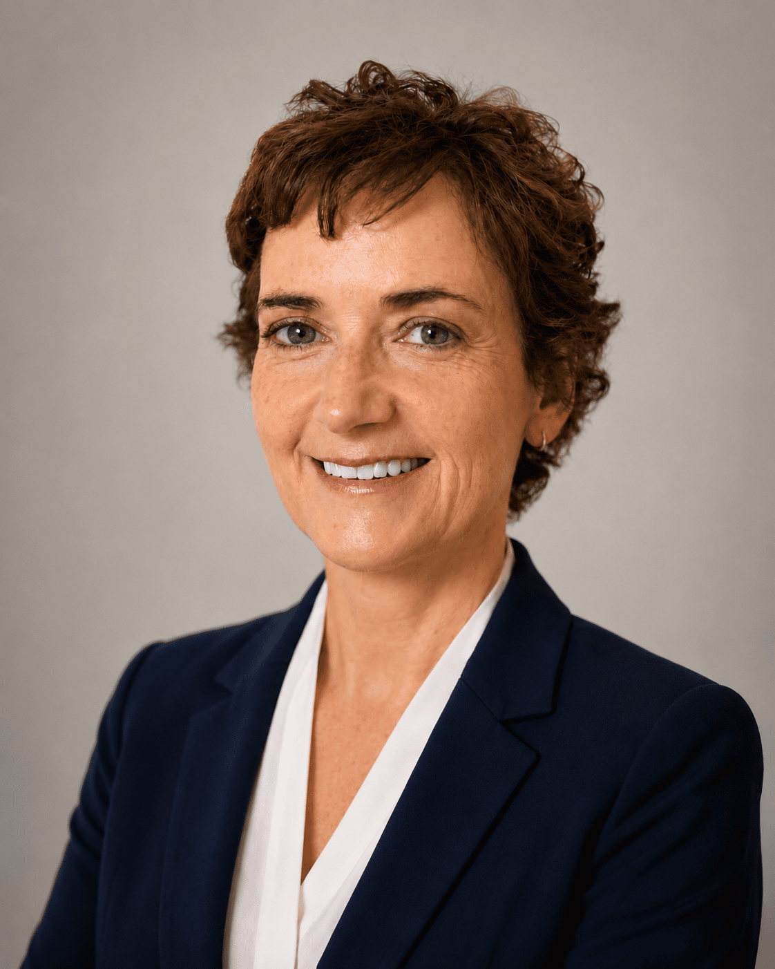 Surgeon Siobhan O’Ceallaigh with short curly brown hair, blue eyes, and light skin, smiling at the camera, wearing a navy blazer and white blouse, against a plain grey background.