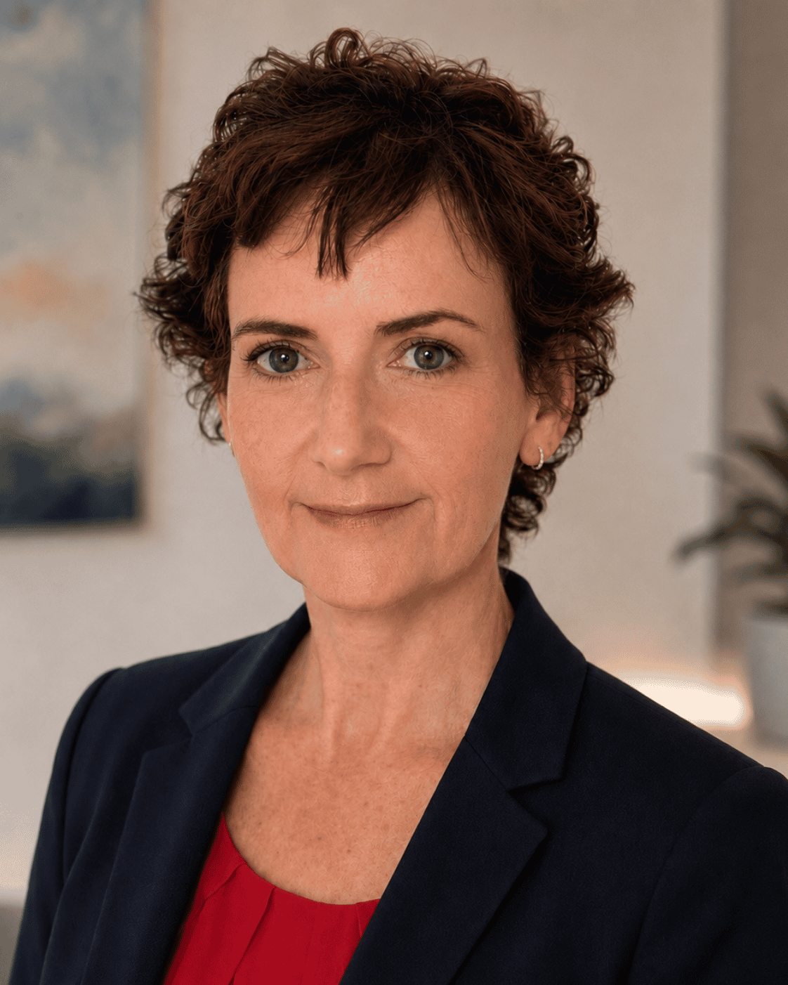 Surgeon Siobhan O’Ceallaigh with short, curly brown hair, wearing a dark blazer over a red blouse, looking at the camera with a neutral expression in an office setting.