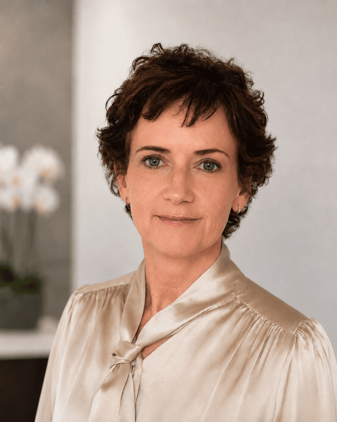 Surgeon Siobhan O’Ceallaigh with short curly brown hair and blue eyes wearing a beige silk blouse with a bow tie, looking confidently at the camera in an indoor setting with blurred background.