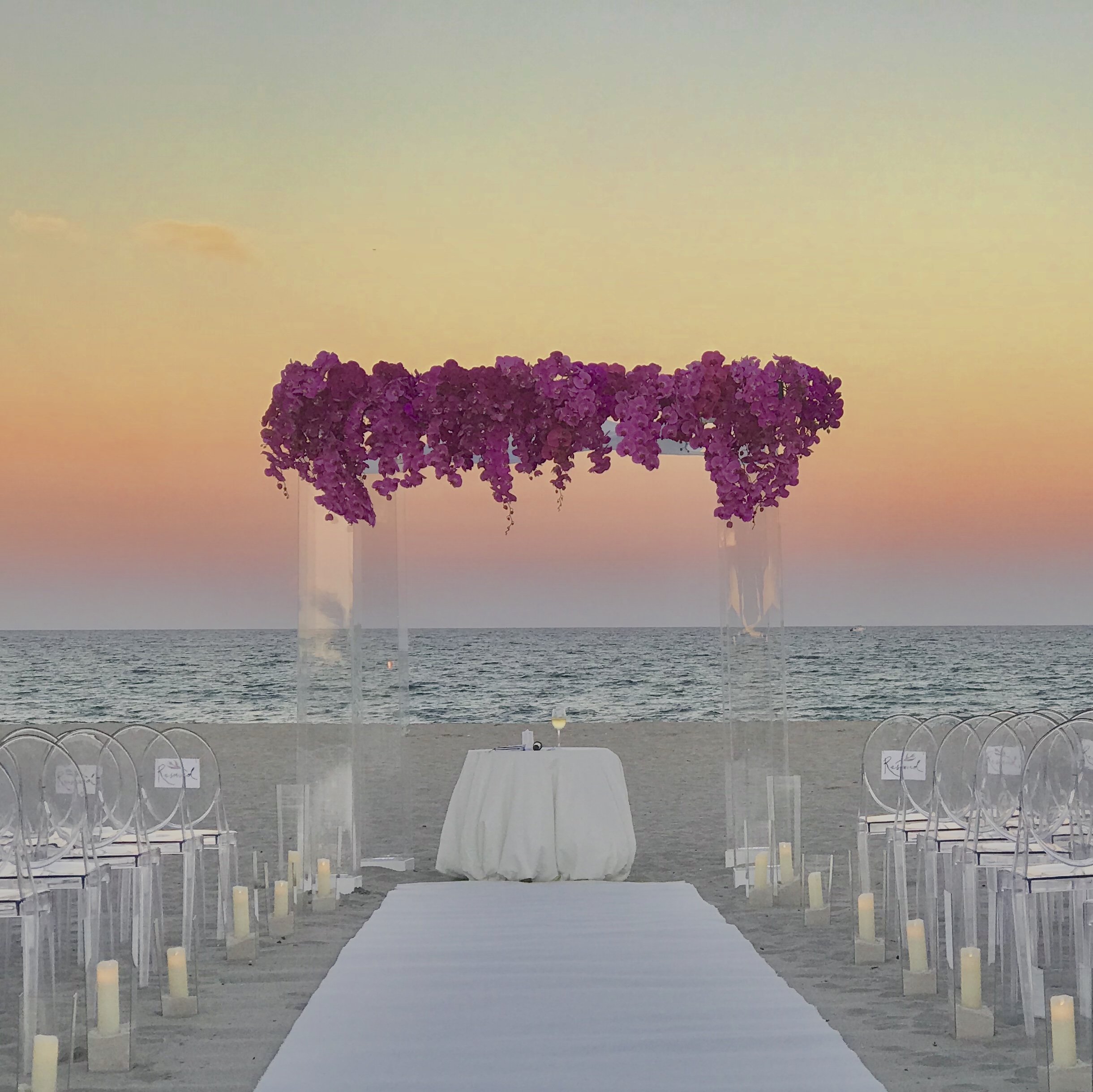 An outdoor wedding ceremony with acrylic arch setup by the beach at sunset, with chairs arranged along a white aisle and small candles on the ground.