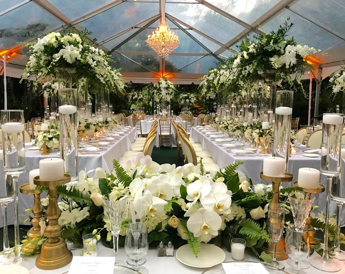 Elegant wedding reception setup with long banquet tables decorated with white floral arrangements, candles in tall glass holders, and gold accents. The tent features a glass ceiling with chandelier lighting.