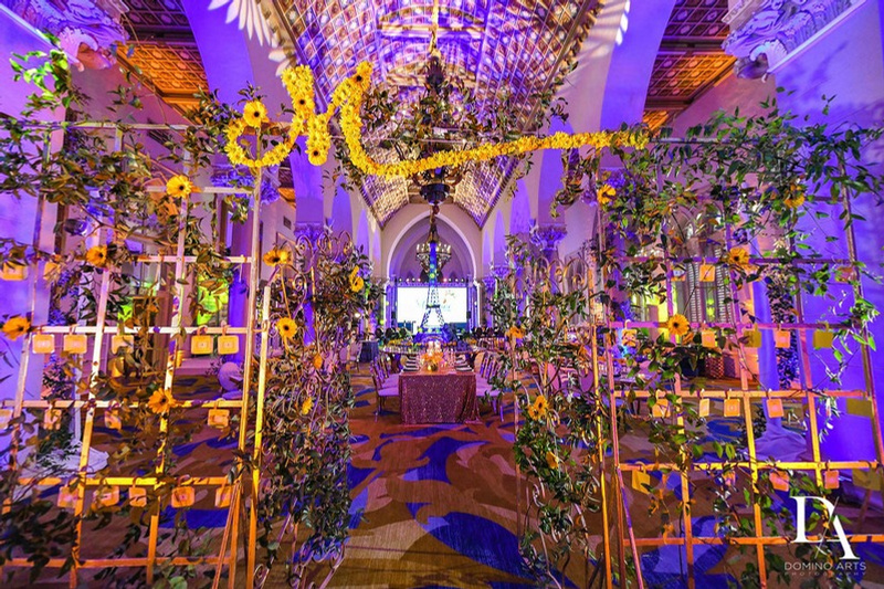 Decorated event hall with arched ceilings, chandeliers, sunflower and greenery centerpieces, and purple lighting.