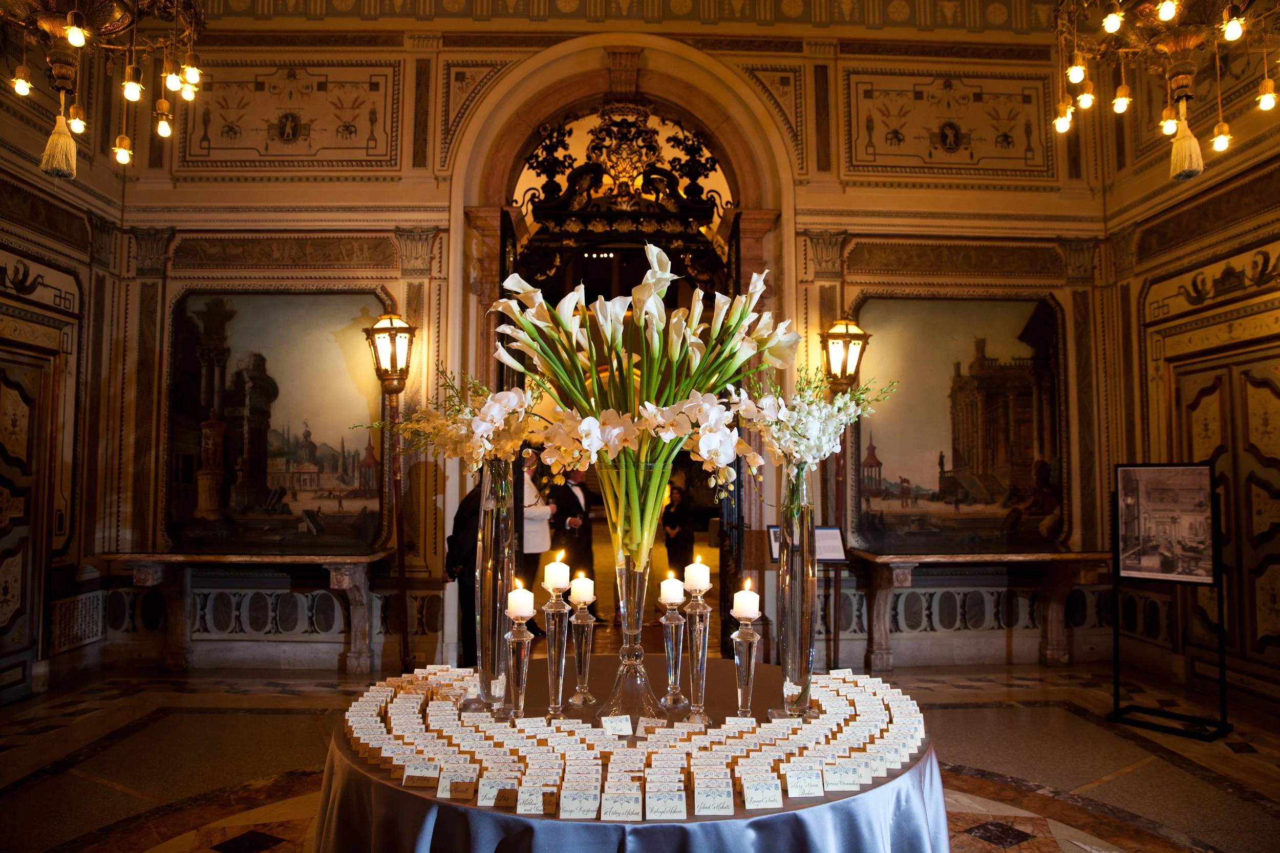 Elegant indoor setting with a table of white flowers in tall vases, surrounded by small name cards, in an ornately decorated room with paintings, chandeliers, and intricate moldings.