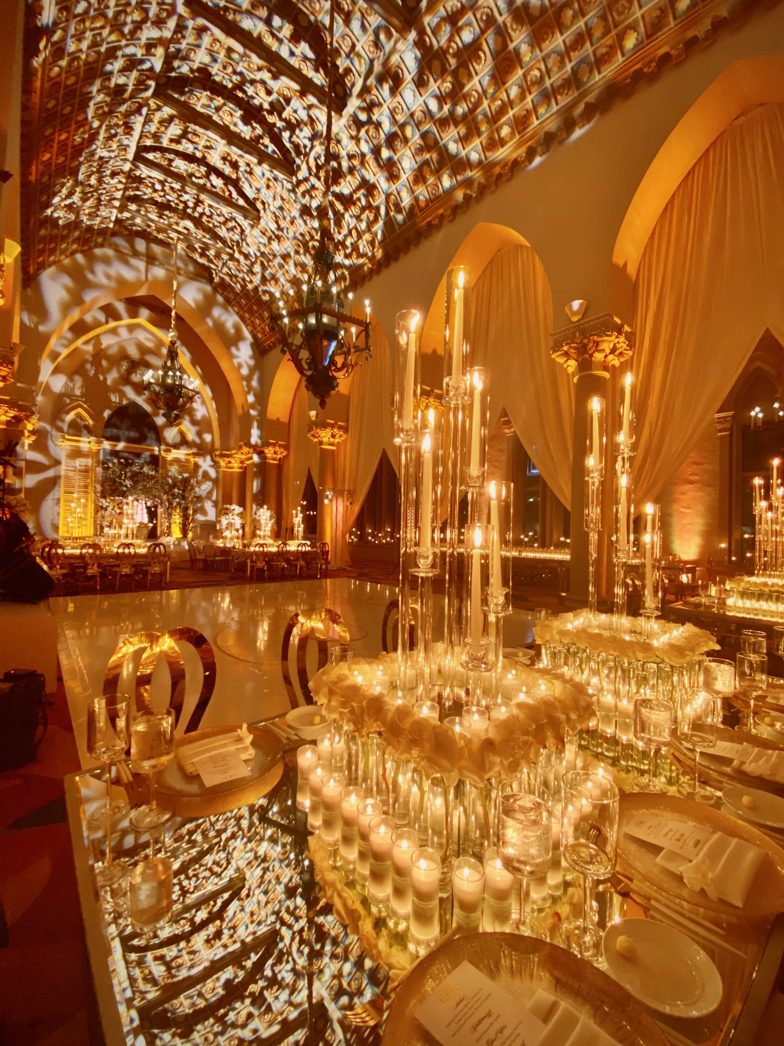 Elegant banquet hall decorated for a celebration with warm amber lighting, candles, tall floral arrangements, and gold accents. The ceiling features intricate artwork and lighting projections, creating a luxurious atmosphere.