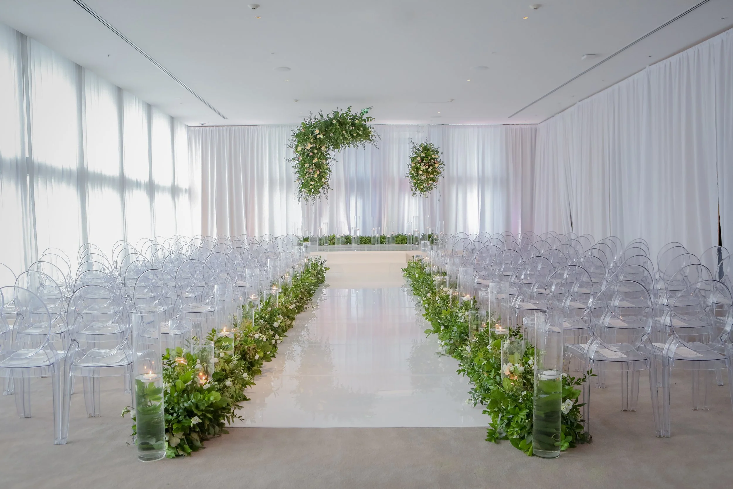 Empty wedding ceremony with clear chairs and floral arrangements along the aisle in a bright, modern venue.
