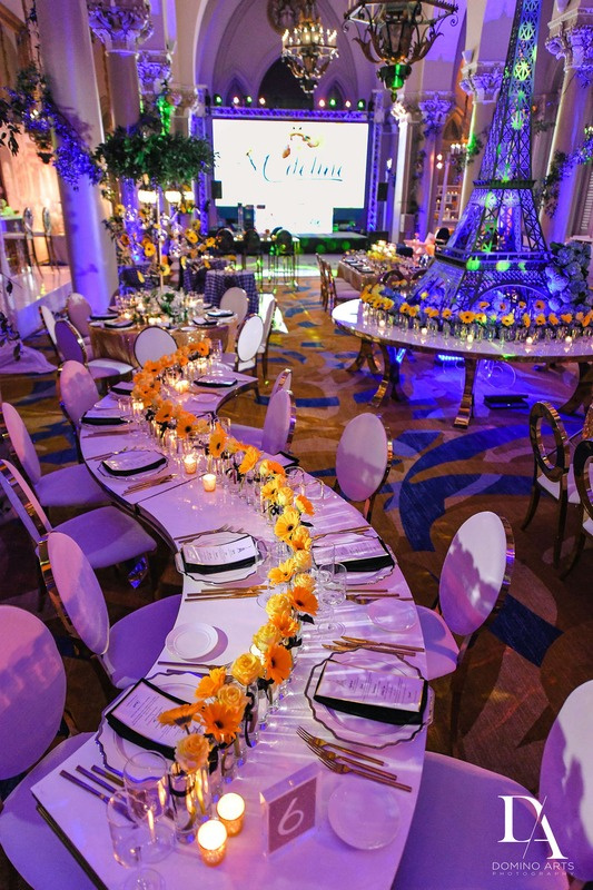 Elegant banquet hall decorated for a celebration with a long floral centerpiece of yellow flowers, candles, and lit table settings, a stage with a large screen, and whimsical decor including a miniature Eiffel Tower adorned with flowers.