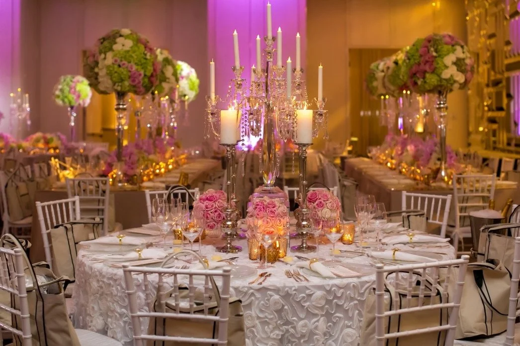 Elegant banquet table with a white embroidered tablecloth, pink rose centerpieces, tall candleholders with lit white candles, crystal chandeliers, and floral arrangements in the background, set for a formal event or wedding reception.