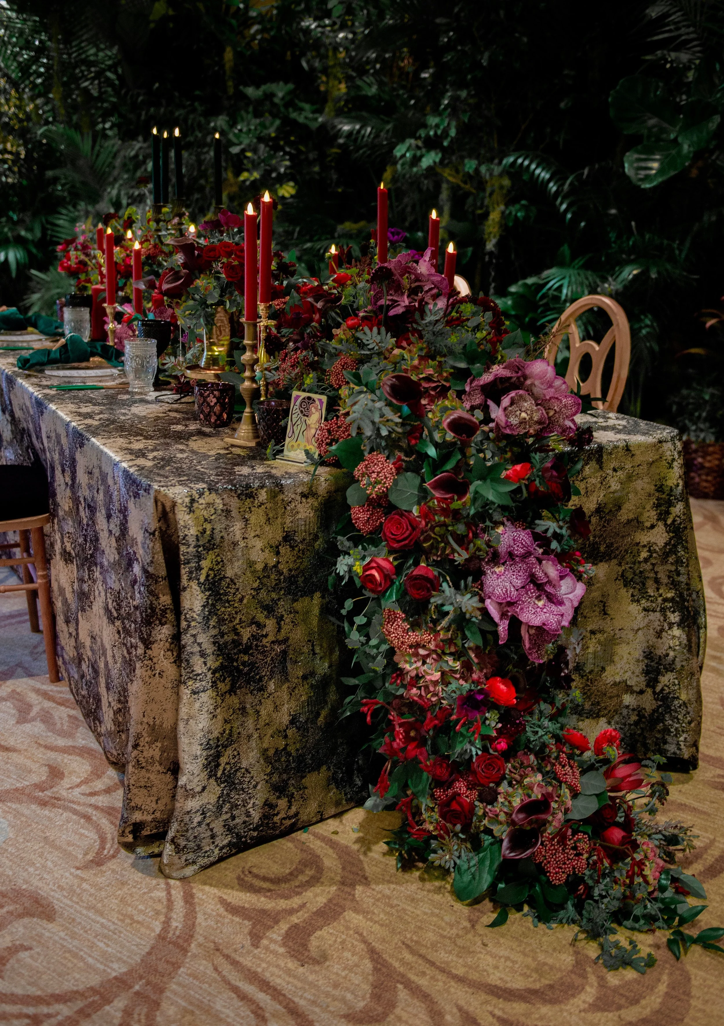 A decorated table with red and pink flowers, candles, and drinkware, set against a dark leafy background in a garden or indoor venue.