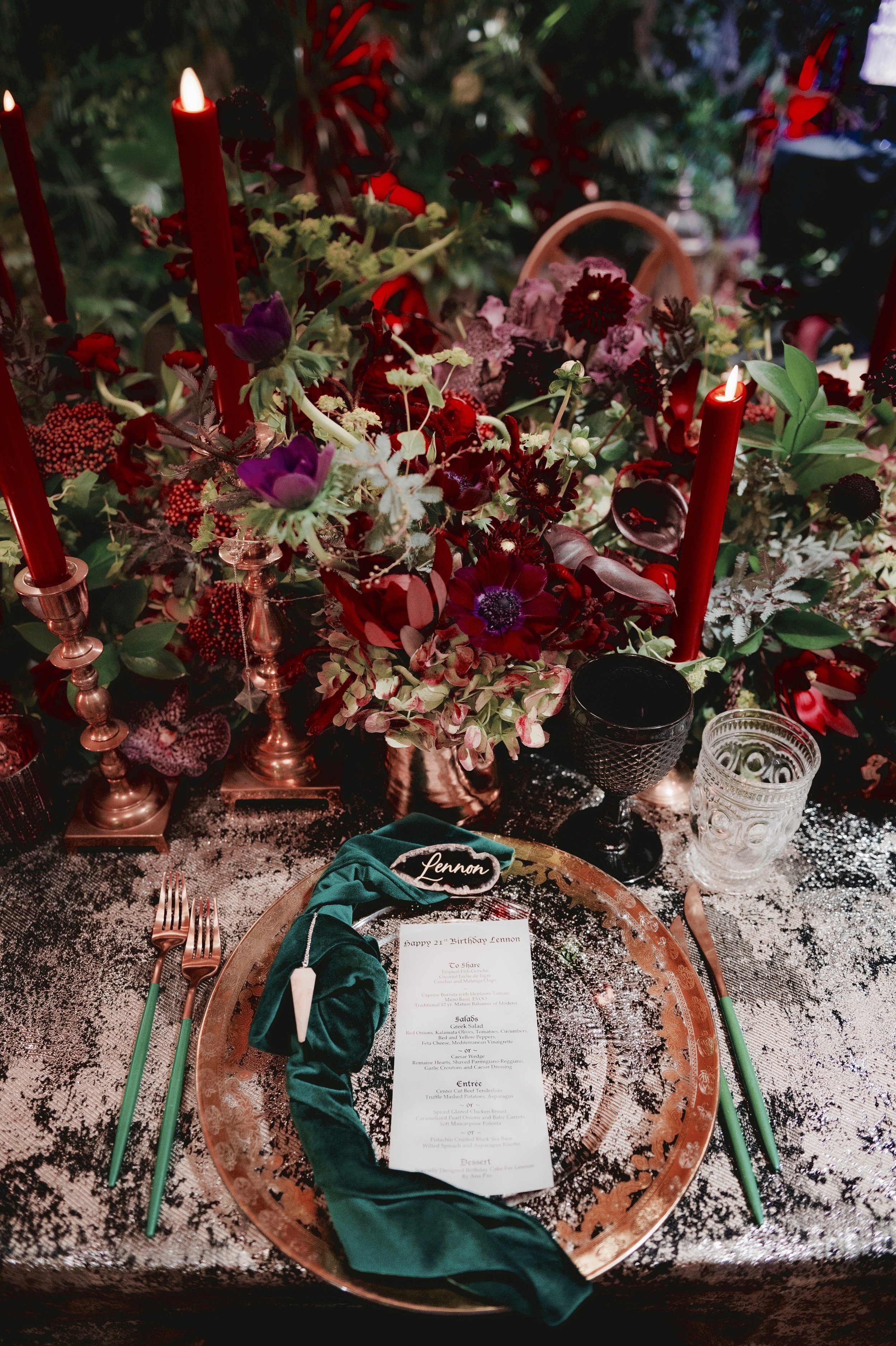 A festive dinner table setting featuring a large bouquet of red, purple, and white flowers, red candles, and elegant glassware. The table has a silver and black patterned tablecloth, a menu card, and a green napkin with a name tag reading 'Lennon' pl