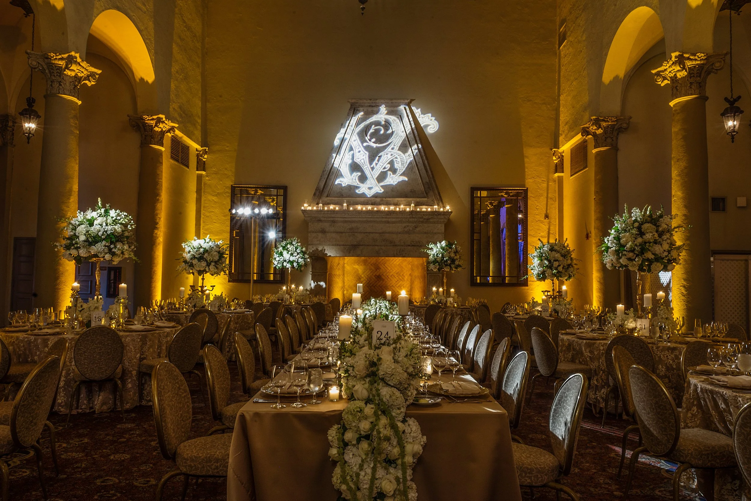 Elegant banquet hall decorated with white floral centerpieces and candles on round tables, illuminated by warm yellow lighting, with a large illuminated crest or monogram on the back wall.