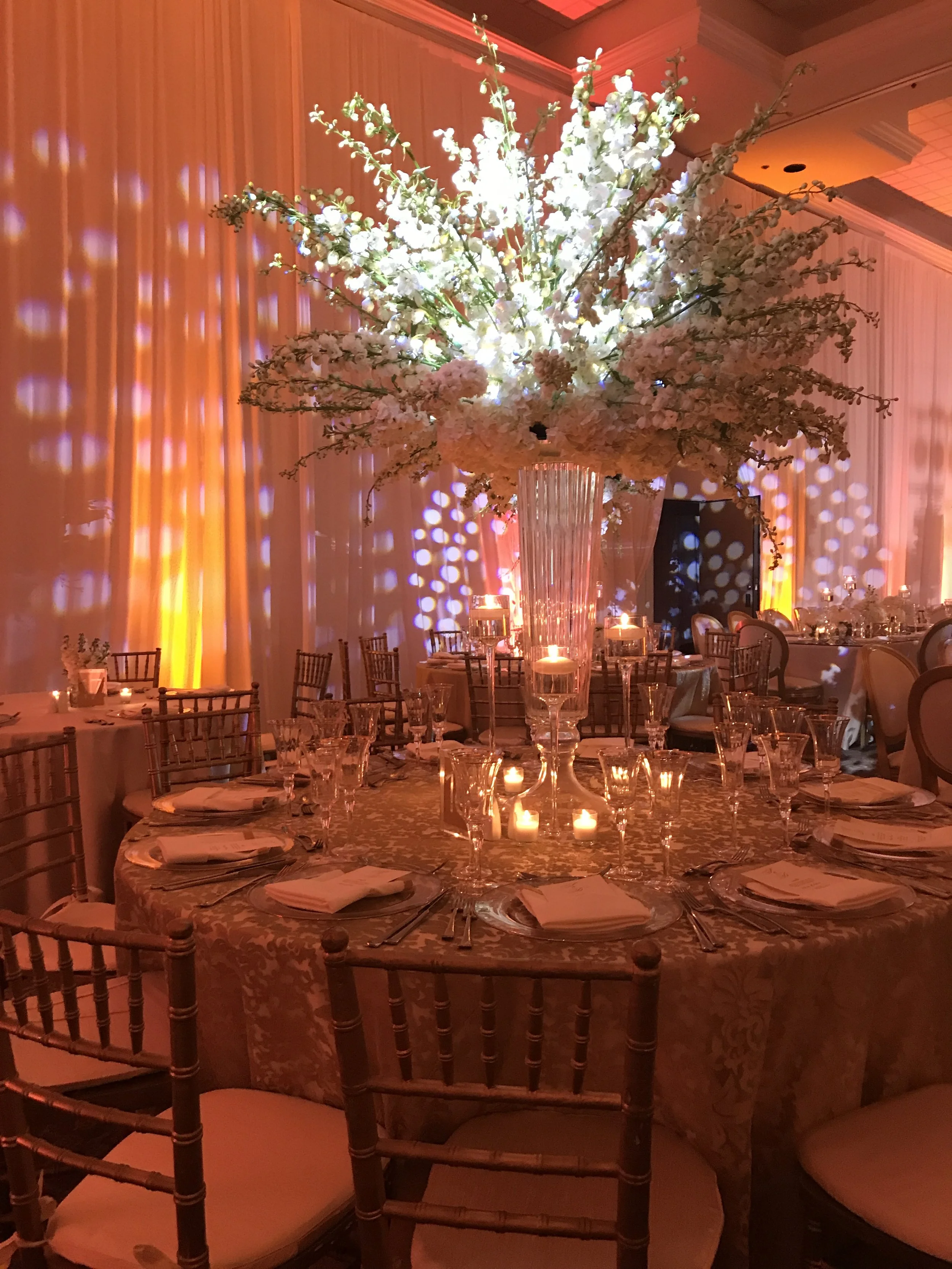 A round dining table with a floral tablecloth set for a formal dinner, surrounded by chairs, with tall candleholders and a large floral centerpiece at the center, in a dimly lit banquet hall with warm orange lighting.