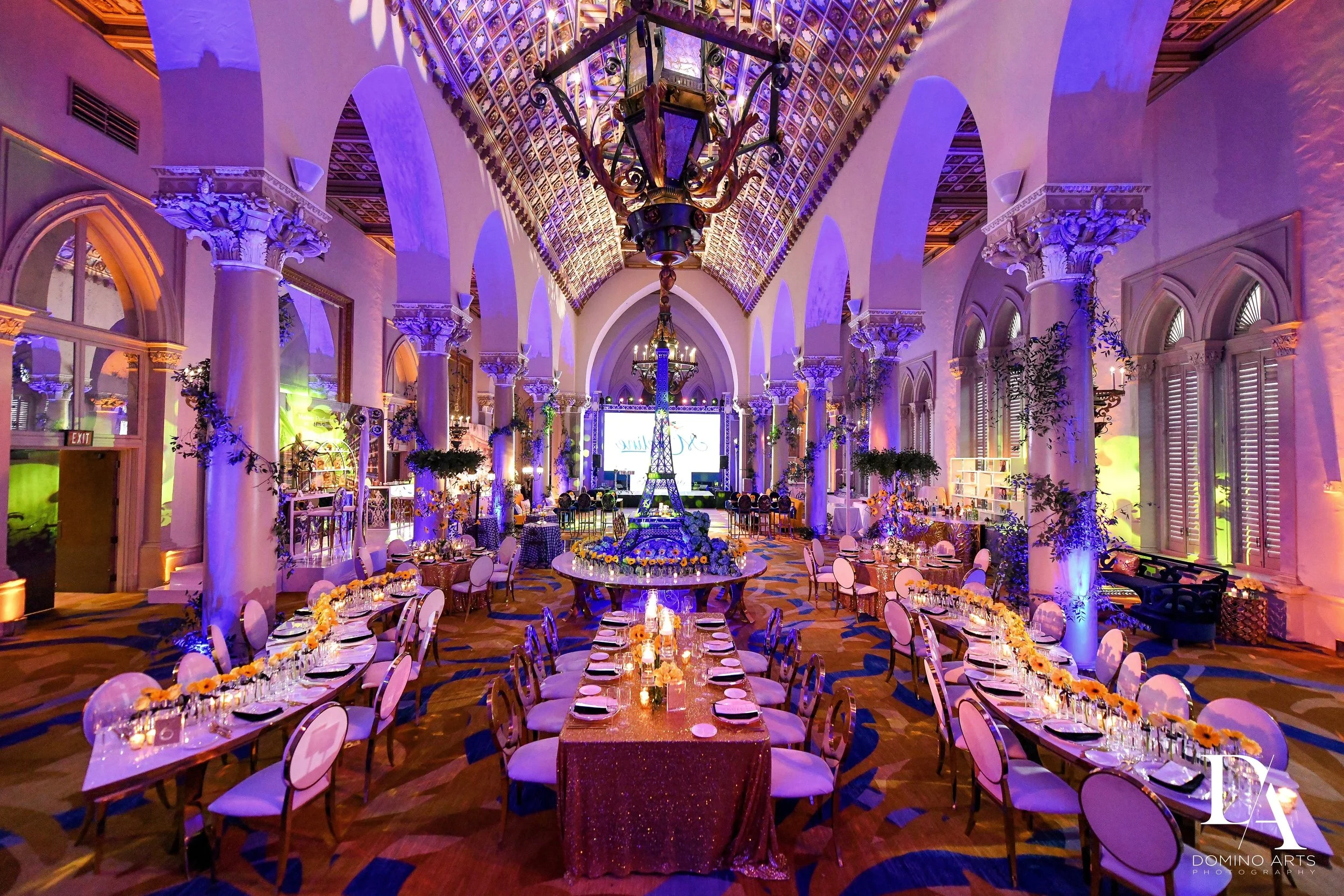A banquet hall decorated for a celebration with tables covered in gold glittering tablecloths and floral centerpieces, purple lighting illuminating the room, ornate white columns with decorative capitals, a vaulted ceiling with intricate design, chan