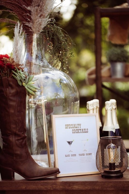 A display with a large glass vase filled with pampas grass and greenery, a pair of champagne bottles, a framed sign for specialty cocktails, and a decorative lantern on a wooden surface, with plants and outdoor furniture in the background.