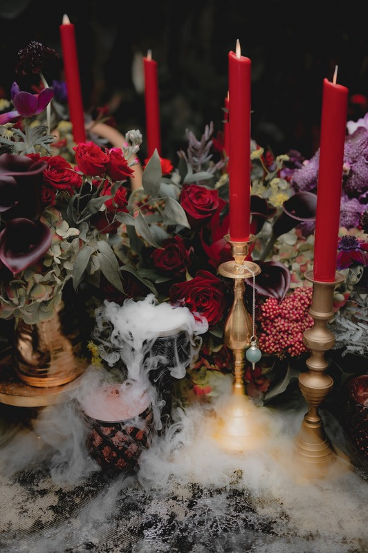 A table decorated with red candles, red roses, purple flowers, and foliage, with a smoky effect in the foreground.