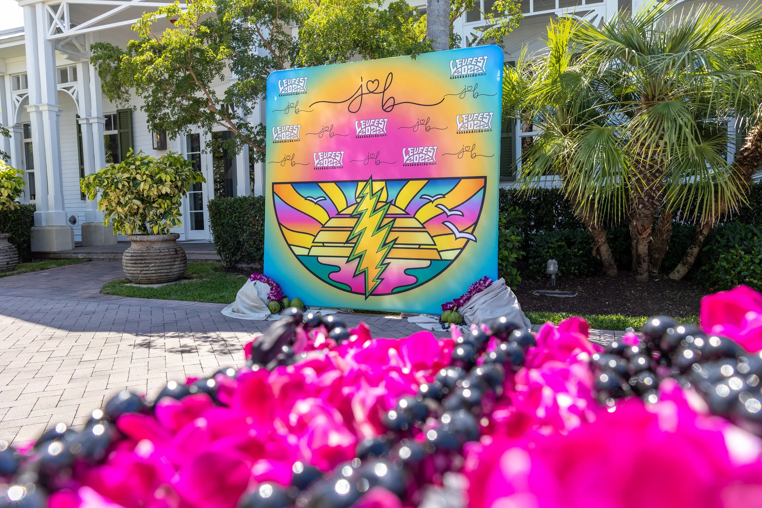 Colorful sign with text and graphics during a festival, with lush green plants and a white building in the background.