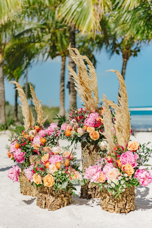 Floral arrangements with pink, orange, and white flowers on bark pedestals, with tall pampas grass, placed on a sandy beach with palm trees and blue ocean in the background.